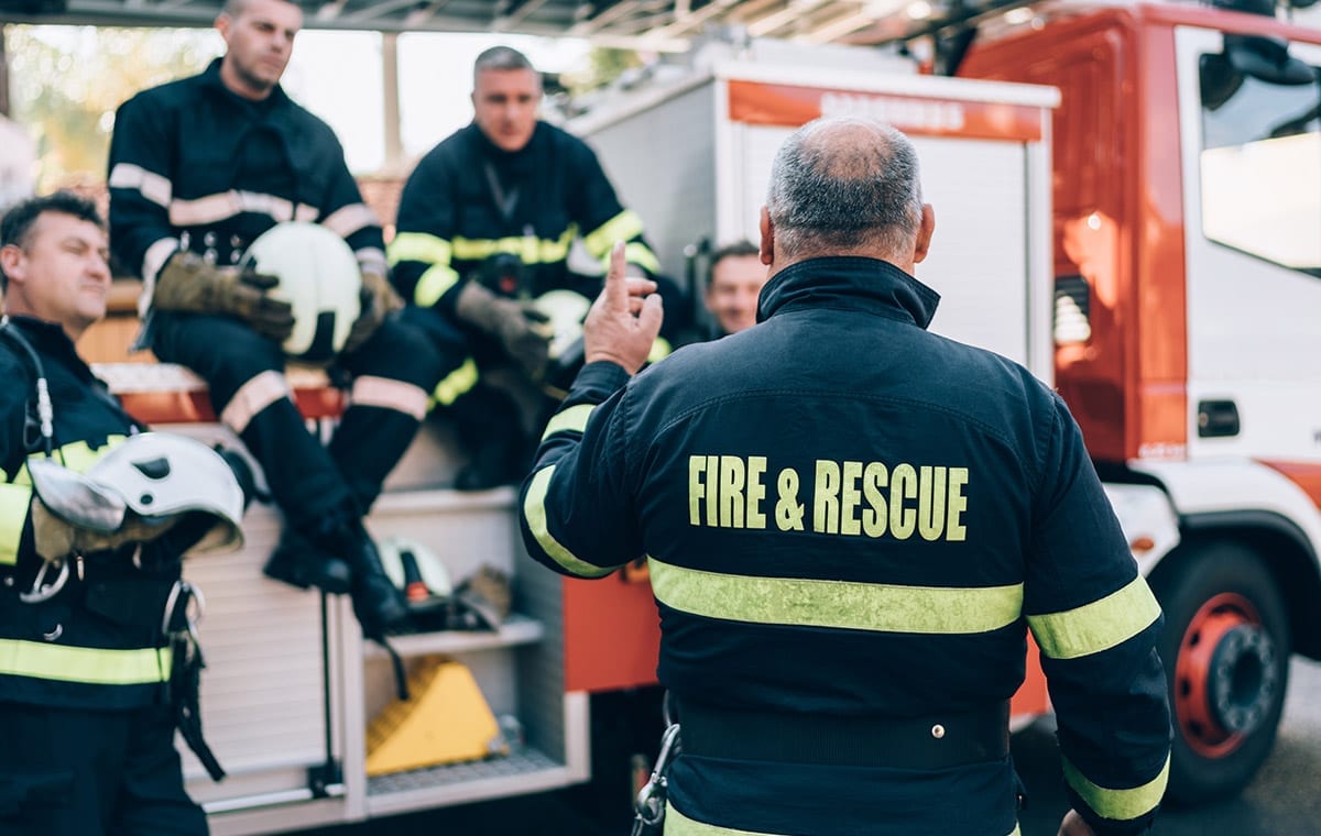 Image of a group of fireman sitting by their firetruck and having a discussion.